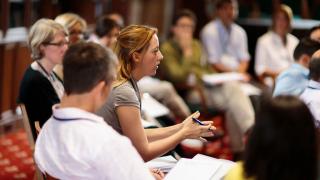 Woman in grey t-shirt talking in front of a group of listeners.